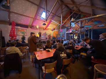 Ein lebendiger Veranstaltungsort mit farbenfroher Dekoration, Menschen, die an Tischen sitzen, und einer Person, die auf der Bühne spielt. Das Publikum ist aufmerksam und schafft eine gemütliche, lebendige Atmosphäre.