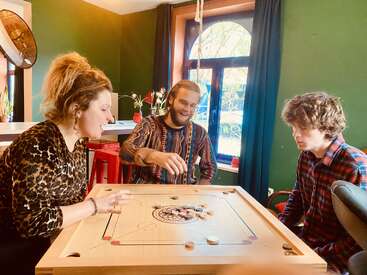 Drei Personen spielen drinnen eine Partie Carrom. Sie sitzen um ein hölzernes Carrom-Brett herum, lächeln und genießen ihre gemeinsame Zeit in einer gemütlichen Umgebung.