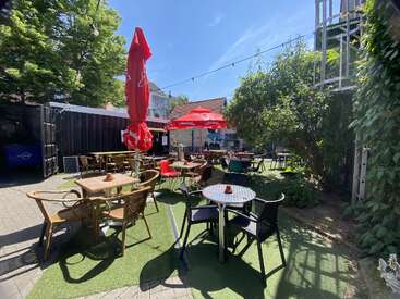 This outdoor cafe features various tables and chairs, red umbrellas, artificial grass, string lights overhead, and is surrounded by lush greenery on a sunny day.
