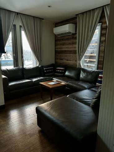 Dieses gemütliche Wohnzimmer verfügt über ein dunkles Ledersofa, einen Couchtisch aus Holz, Zierkissen, Holzböden, große Fenster mit Vorhängen und eine an der Wand montierte Klimaanlage.
