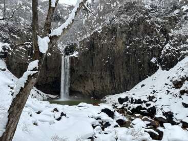 Eine malerische Winterlandschaft mit einem hohen Wasserfall, der in ein Becken stürzt, umgeben von schneebedeckten Felsen, Bäumen und schroffen Klippen unter einem kalten, grauen Himmel.