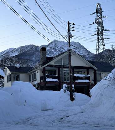 Ein Haus steht inmitten von tiefem Schnee, im Hintergrund sind Berge und Stromleitungen zu sehen. Die winterliche Szenerie wirkt friedlich, klar und eingebettet in die Natur.