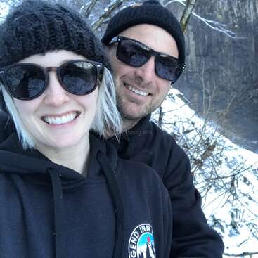 Ein lächelndes Paar in schwarzen Kapuzenpullis und Sonnenbrillen posiert draußen für ein Selfie. Verschneite Bäume und Felsen bilden hinter ihnen eine wunderschöne, winterliche Kulisse.