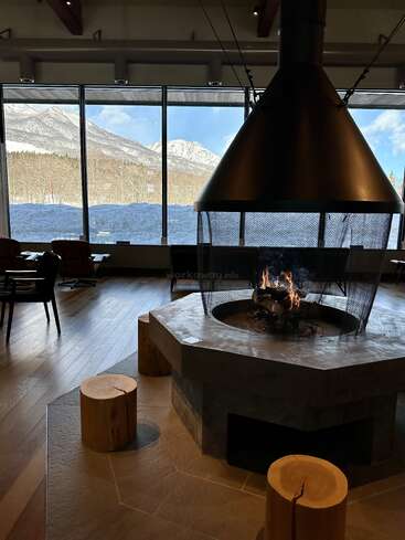 Eine gemütliche Lounge mit einem Kamin in der Mitte, Holzhockern und Stühlen. Durch die großen Fenster blickt man auf schneebedeckte Berge und Bäume, was im Innenraum eine warme, einladende Atmosphäre schafft.