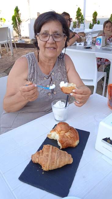 A smiling woman sits at an outdoor café, enjoying a pastry with cream. In front of her are a croissant, brioche, and a glass of dessert.