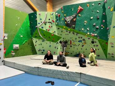 Quatre personnes sont assises sur des tapis rembourrés dans une salle d'escalade intérieure colorée, tandis qu'une personne escalade le mur derrière elles. Des prises lumineuses décorent le mur.