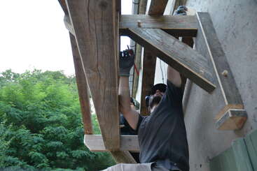 Une personne portant des gants et un bonnet travaille sur une structure en bois fixée à un mur, en utilisant des outils et en apparaissant pour l'assembler ou la réparer à l'extérieur.