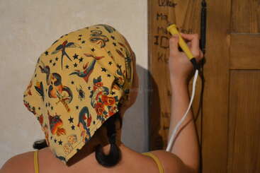 Une personne aux cheveux tressés et au foulard coloré utilise un outil jaune et noir pour écrire ou graver sur une surface en bois.