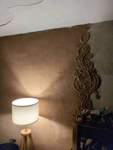In einer gemütlich beleuchteten Zimmerecke gibt es eine kunstvoll gestaltete, strukturierte Wand, eine Stehlampe auf drei Beinen, eine weiß getünchte Decke mit Rissen und einen kleinen, vollgestopften Tisch, auf dem allerlei Krimskrams liegt.