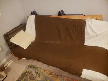 La imagen muestra un sofá futón con funda marrón, almohadas blancas y cabecero de madera, situado en un rincón de una habitación con una alfombra en el suelo.