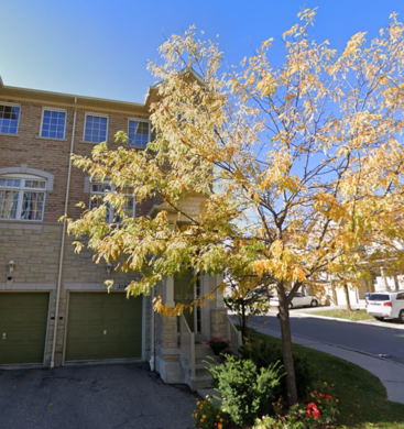 La imagen muestra una casa adosada de dos plantas con una puerta de garaje verde, un árbol de hojas amarillas en primer plano y un sereno cielo azul en lo alto.