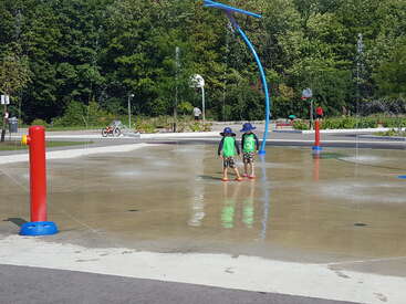 La imagen muestra a dos niños en un chapoteadero, con camisetas y gorros verdes, de pie en una zona de hormigón con fuentes de agua y árboles al fondo.
