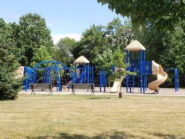 La imagen muestra un parque infantil de color azul intenso con tejados de color tostado, que incluye un tobogán, una estructura para trepar y barras para montar, rodeado de árboles y una zona de césped en un día soleado.