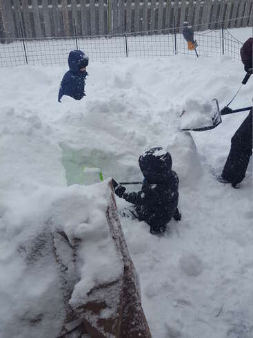 La imagen representa un patio trasero nevado con niños jugando en la nieve, una pala y una valla al fondo, evocando una sensación de diversión y actividad invernales.