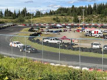 The image depicts a race track with a white car on the track, surrounded by a parking lot filled with cars and trucks, and a grassy hill in the background.