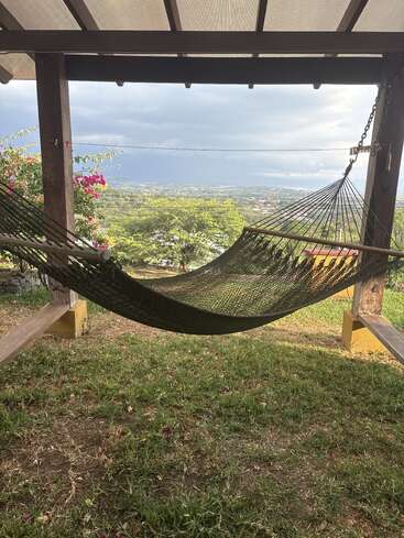Un hamac confortable est suspendu sous une structure en bois ombragée, donnant sur un paysage pittoresque composé d'arbres verts, de montagnes lointaines et d'un ciel nuageux, créant ainsi un havre de paix.