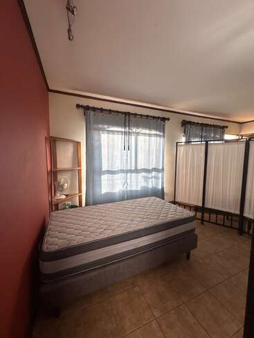 Une chambre sobre, avec un matelas nu, un mur d'accent rouge, des rideaux bleus, un sol carrelé, une petite étagère surmontée d'un ventilateur et un paravent pliant. La lumière du soleil y pénètre.