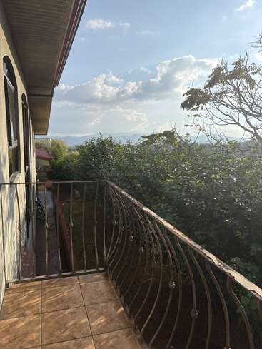 Un petit balcon carrelé, doté d'une balustrade métallique, donne sur des arbres verdoyants. Le ciel est clair, parsemé de nuages, et on aperçoit les montagnes au loin.