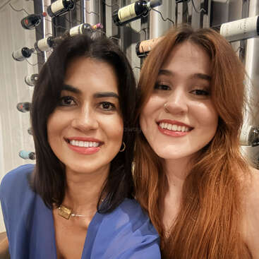 Deux femmes sourient ensemble pour un selfie devant un casier à bouteilles. Elles ont toutes deux l'air heureuses et détendues, avec des bouteilles de vin disposées derrière elles.