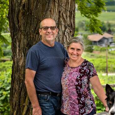 A smiling couple stands close together in front of a large tree, with lush greenery and a house in the background. Part of a dog appears.