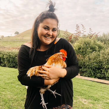 Uma mulher com longos cabelos escuros, vestindo uma camisa preta, segura um galo nos braços em um campo gramado com arbustos e uma colina ao fundo.
