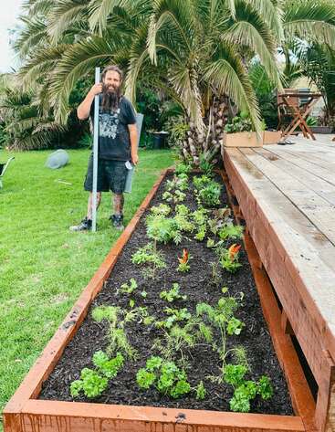 A imagem mostra um homem em pé ao lado de um canteiro de jardim elevado, cercado por uma vegetação exuberante e palmeiras, em um ambiente externo sereno.