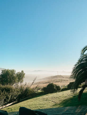 A imagem retrata uma paisagem serena com uma colina gramada, palmeiras e um deck de madeira, em um céu azul claro e um horizonte distante e nebuloso.