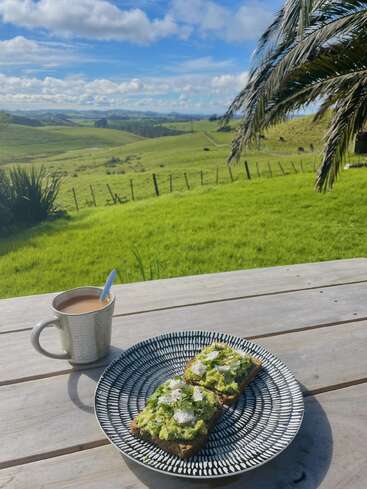 A imagem retrata um ambiente externo sereno com um prato de torrada de abacate, uma caneca de café e uma paisagem verde pitoresca sob um céu azul com nuvens brancas.