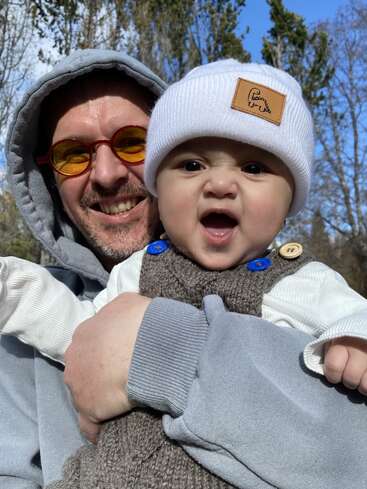 Un homme souriant portant des lunettes et un sweat à capuche tient dans ses bras un bébé portant un bonnet blanc et une salopette marron, en plein air par une journée ensoleillée au milieu des arbres.