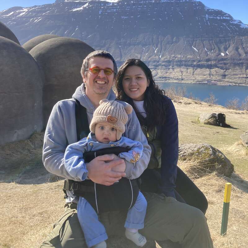 Une famille souriante de trois personnes pose en plein air ; le père tient son bébé dans un porte-bébé, la mère est assise à côté de lui. Des montagnes, de l'eau et des structures en forme de dôme créent un arrière-plan pittoresque.