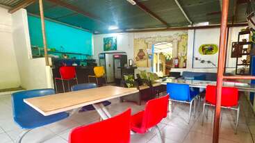 Dieses Bild zeigt ein gemütliches, legeres Café mit bunten Plastikstühlen, Holztischen, einem kleinen Küchenbereich, Wandkunst, Sofas und einer hellen, einladenden Einrichtung.