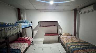 Dieses Bild zeigt ein kleines, einfaches Zimmer mit drei Einzelbetten, gefliestem Boden, weißen Wänden, einem Regal, einer Klimaanlage und wenig natürlichem Licht von oben.