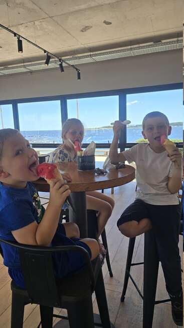 Trois enfants dégustent des glaces colorées à une table dans un café moderne doté de grandes fenêtres donnant sur un front de mer pittoresque et ensoleillé et un ciel bleu à l'extérieur.