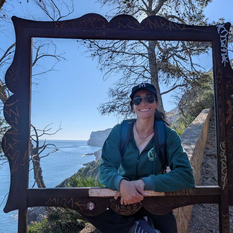 Une femme souriante, portant des lunettes de soleil et une casquette, est appuyée contre un cadre décoratif, avec en arrière-plan une vue panoramique sur la côte, des arbres, un ciel bleu et des falaises.