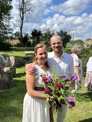 Ein lächelndes Paar steht an einem sonnigen Tag im Freien. Die Frau hält einen bunten Blumenstrauß in der Hand. Im Hintergrund sind andere Menschen und üppiges Grün zu sehen.