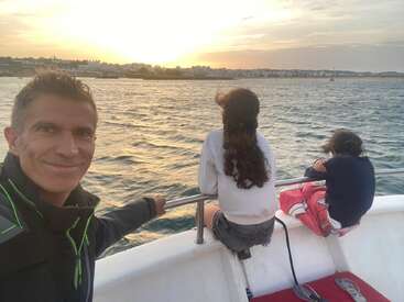 Un hombre sonríe a la cámara en un barco al atardecer, mientras dos niños sentados en la barandilla contemplan las aguas tranquilas y la costa lejana.