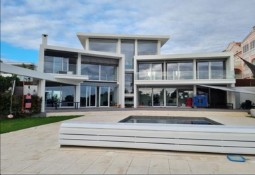 Una casa moderna de tres plantas con grandes ventanales de cristal, tejados planos, arquitectura angulosa y blanca, un patio espacioso, una pequeña piscina y toques de vegetación.