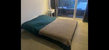 Un dormitorio minimalista con una cama baja con fundas verdes y beige, una mesilla de noche blanca, suelo de baldosas y grandes puertas de cristal que dan a un patio con asientos al aire libre.