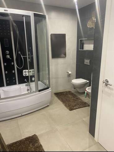 Este moderno cuarto de baño cuenta con una cabina de ducha de cristal, inodoro montado en la pared, bidé, alfombras marrones, azulejos grises, un espejo rectangular y un mínimo de accesorios decorativos para lograr un aspecto limpio.