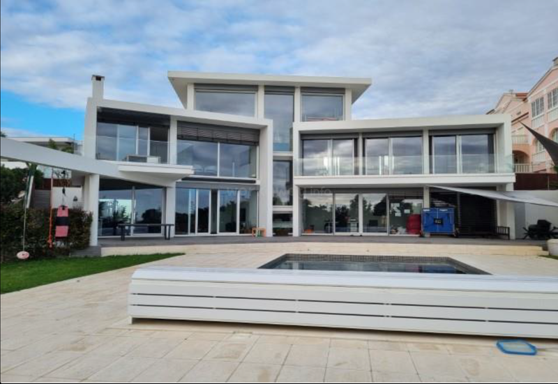 Una casa moderna de tres plantas con grandes ventanales de cristal, tejados planos, arquitectura angulosa y blanca, un patio espacioso, una pequeña piscina y toques de vegetación.