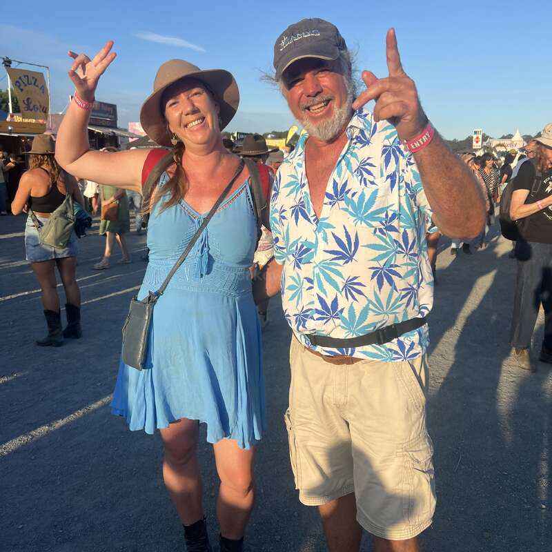Un homme et une femme souriants posent lors d'un festival en plein air, portant des vêtements d'été décontractés et des chapeaux, entourés de gens. L'atmosphère semble joyeuse et détendue.