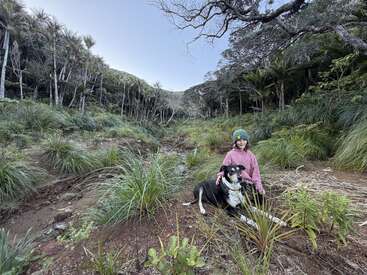 Uma criança com uma jaqueta rosa e um gorro verde está sentada numa clareira verdejante da floresta com seu grande cachorro preto e branco, curtindo a natureza juntos.