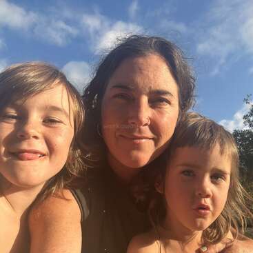 A woman and two children pose closely together outdoors, smiling under clear blue skies. The sunlight highlights their faces, creating a warm and joyful atmosphere.