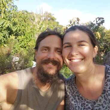 Una pareja sonriente se toma un selfie en primer plano al aire libre en un día soleado, rodeada de frondosos árboles y plantas verdes, irradiando felicidad, amor y calidez.