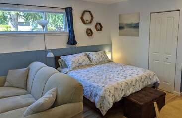 Um quarto aconchegante tem uma cama grande com roupa de cama estampada de flores, um sofá bege, prateleiras hexagonais na parede, quadros com paisagens, luminárias, uma janela e a porta de um armário.