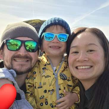 Uma família alegre de três pessoas posa ao ar livre. A criança usa óculos escuros azuis e uma jaqueta amarela, ladeada pelos pais sorridentes. Todos parecem felizes sob um céu ensolarado.
