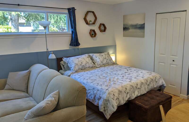 Um quarto aconchegante tem uma cama grande com roupa de cama estampada de flores, um sofá bege, prateleiras hexagonais na parede, quadros com paisagens, luminárias, uma janela e a porta de um armário.