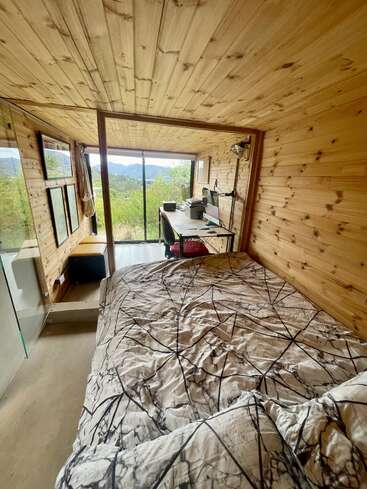 A cozy wooden cabin room with a bed in the foreground, a desk and computer by large windows, offering beautiful views of nature and distant mountains.