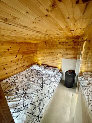 This cozy, compact wooden room features pine walls and ceiling, a neatly made bed with a geometric-patterned duvet, and a small black cylindrical heater in the corner.