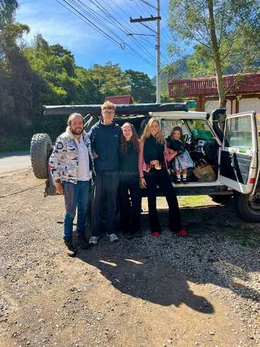 Five people pose happily in front of an off-road vehicle, surrounded by lush greenery and sunlight, with one little girl sitting inside the car’s door.